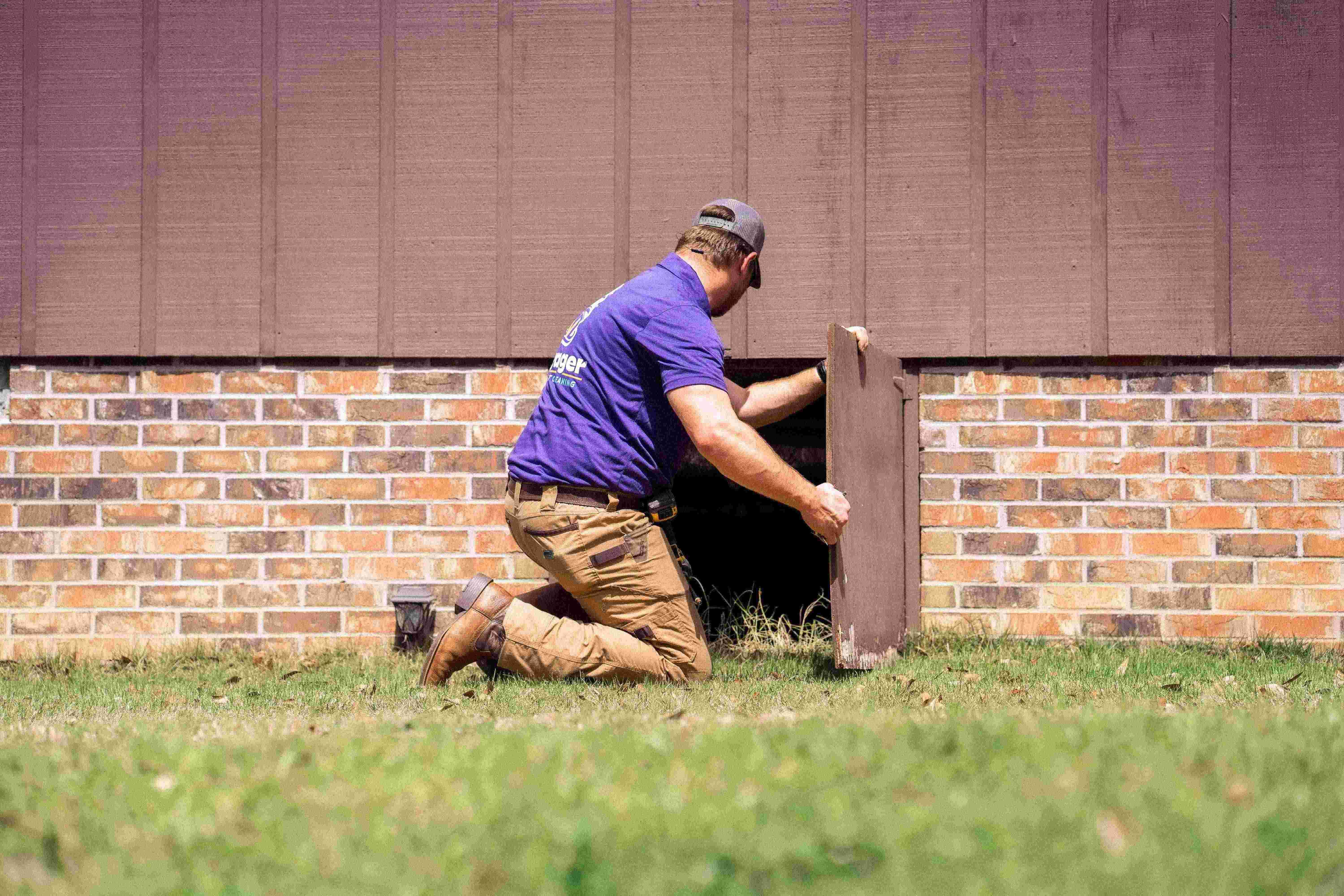 Voyager Air Duct Cleaning specialist kneeling outside a brick home, opening an access panel to reach the crawl space and service the dryer vent.
